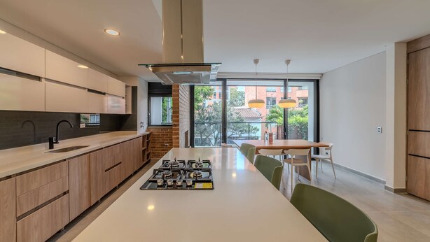 A modern kitchen with a central island, wooden cabinets, and a view of the outdoors through large windows. - w Spacious 3BR w Outdoor Dining Terrace in Laureles (Medellín)