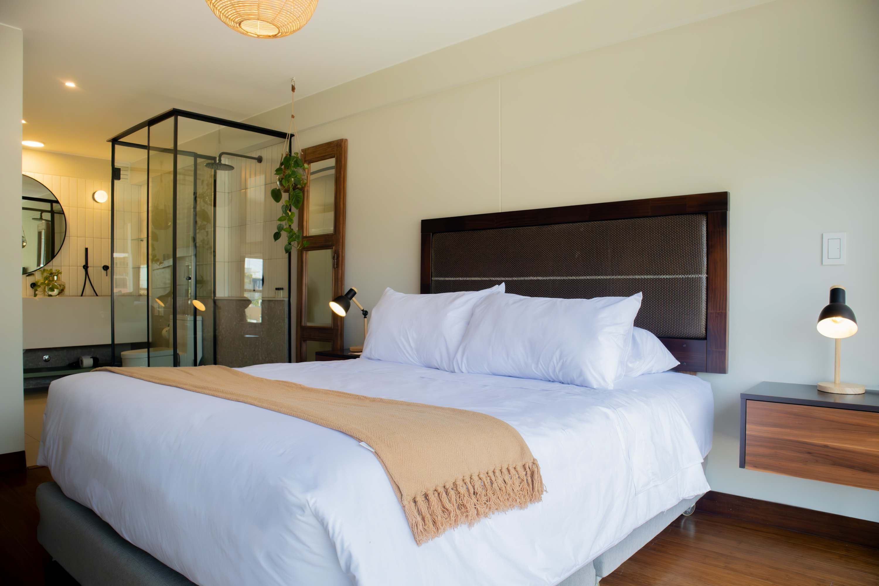 A modern hotel room with a large bed, a glass shower enclosure, a wooden nightstand, and a bedside lamp.