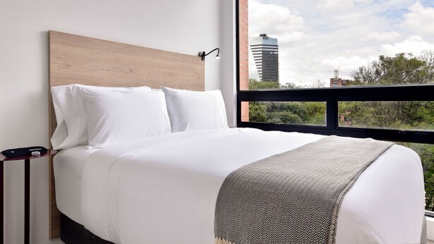 A hotel room with a large bed, a wooden headboard, white bedding, and a view of a cityscape through a large window. - w Stunning Loft in Bogota (Bogotá)