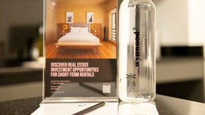 A hotel room with a bed, a nightstand, and a lamp. A bottle of water is placed on the table next to a sign about real estate investment opportunities.