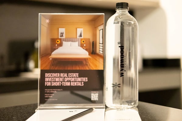 A hotel room with a bed, a nightstand, and a lamp. A bottle of water is placed on the table next to a sign about real estate investment opportunities.