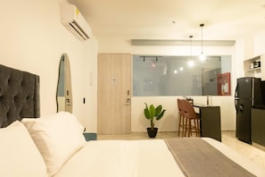 A bedroom with a bed, a desk, a chair, a potted plant, a refrigerator, and a window.
