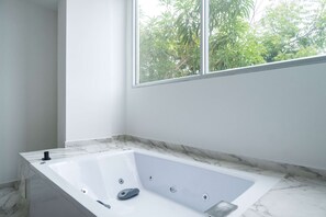 A modern bathroom with a large bathtub, a marble countertop, and a view of greenery through a window.
