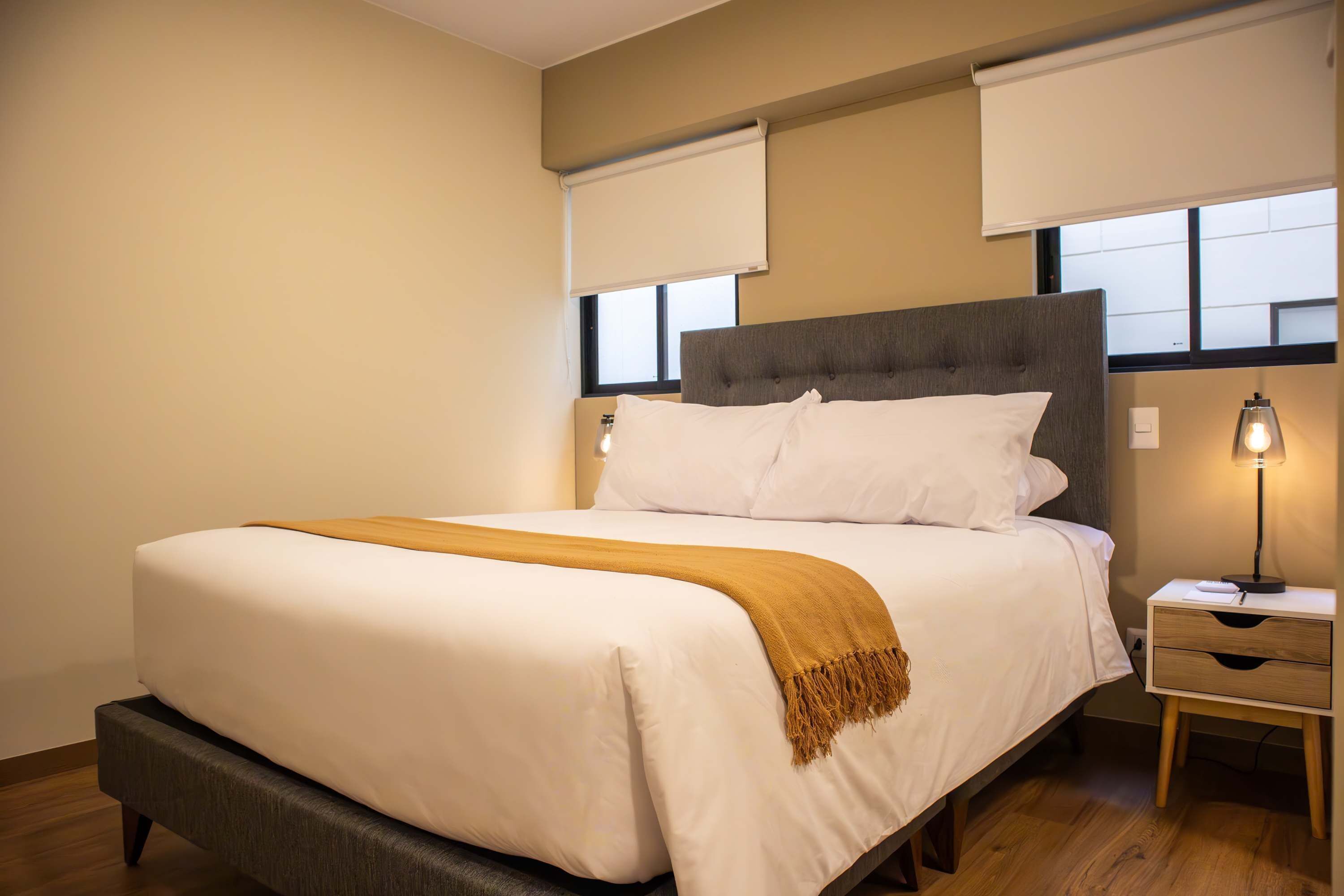 Una cama bien hecha con edredón blanco y manta marrón, una mesita de noche con lámpara, y una ventana con vistas al exterior.