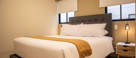 Una cama bien hecha con edredón blanco y manta marrón, una mesita de noche con lámpara, y una ventana con vistas al exterior.