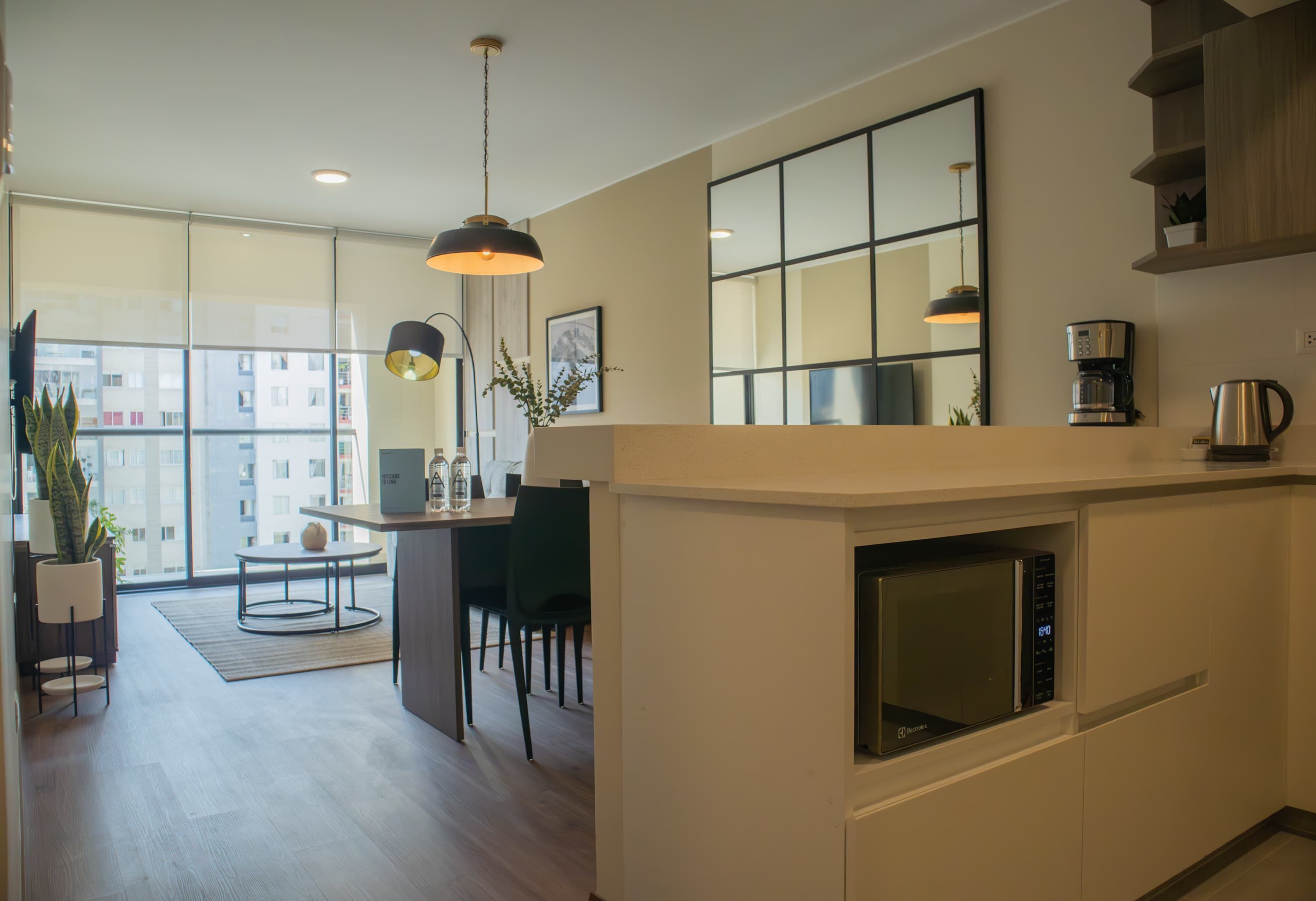 A modern kitchen with a built-in microwave, a coffee maker, and a kettle on the counter. There is a dining area with a table and chairs, and a large window offering a view of the city.