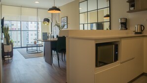 Una cocina moderna con microondas integrado, cafetera y hervidor sobre la encimera. Hay un comedor con mesa y sillas, y un amplio ventanal con vistas a la ciudad.
