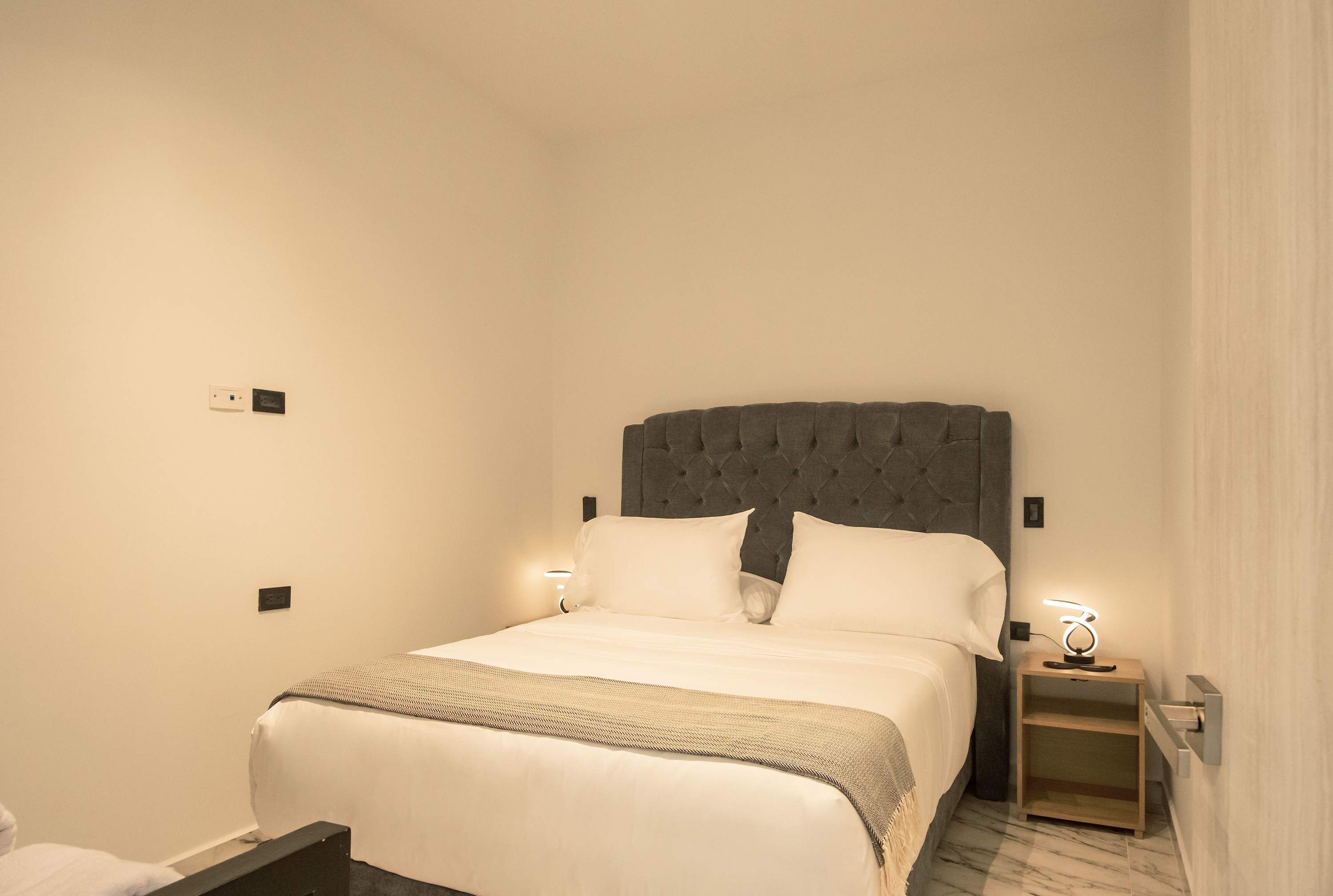 A neatly made bed with a tufted headboard, two bedside tables with lamps, and a marble-patterned floor.