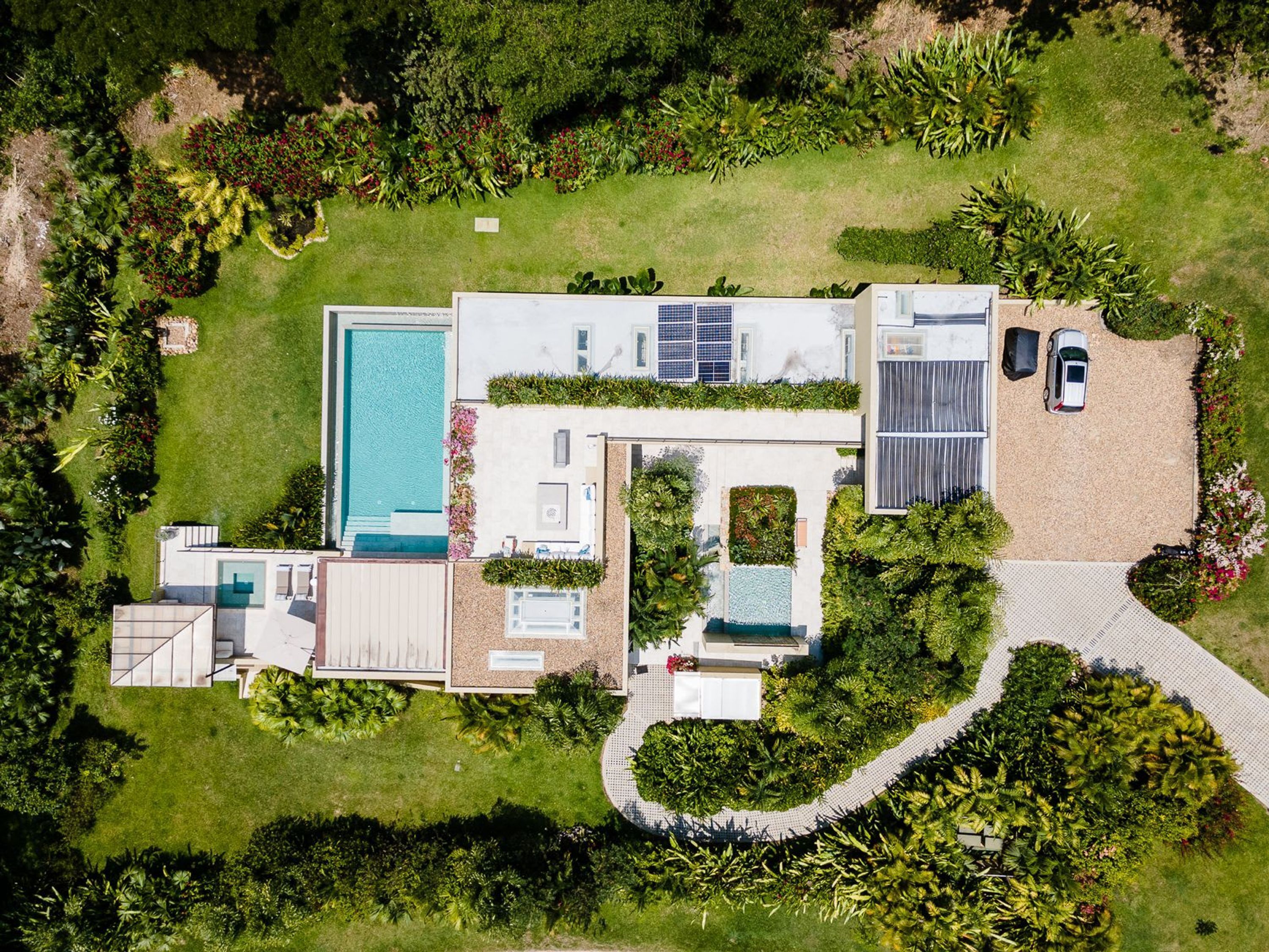 An aerial view of a modern house with a swimming pool, solar panels, and a driveway with two cars.
