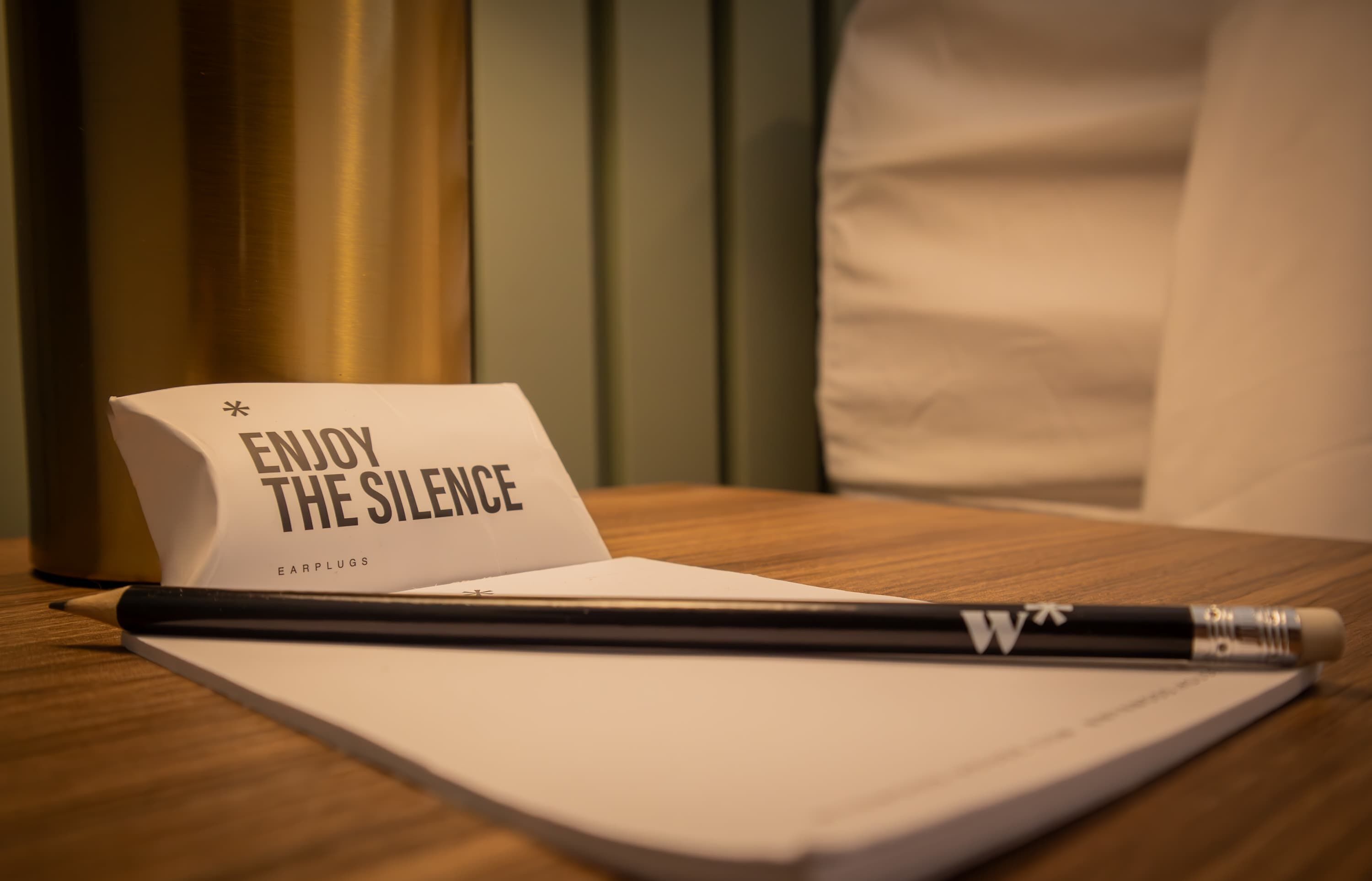 A notebook with a pencil, a card that reads "ENJOY THE SILENCE," and a pair of earplugs on a wooden surface.