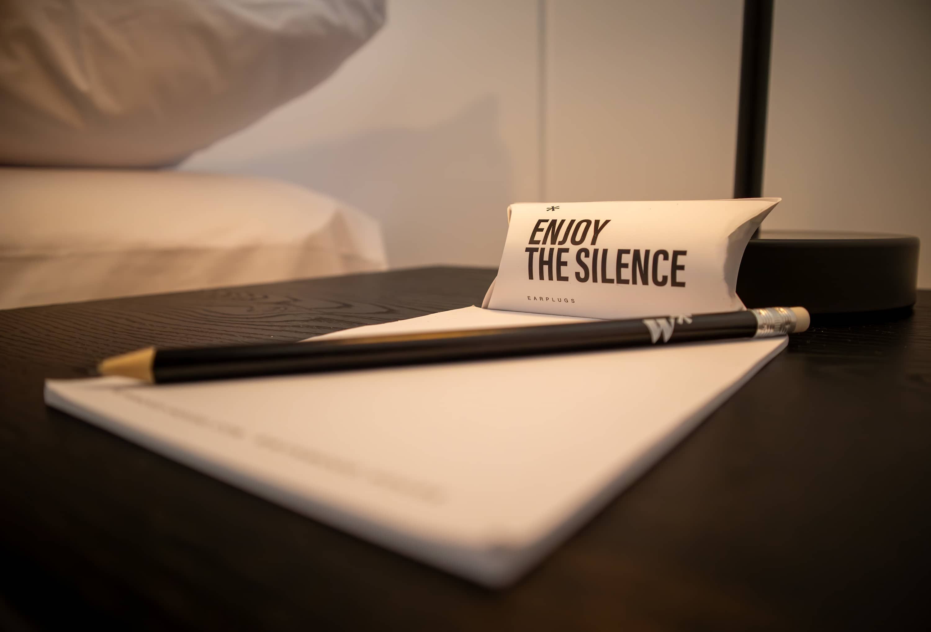 A hotel room with a bed, a bedside table, a laptop, a notepad, and a pen.