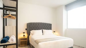 A neatly made bed with a grey upholstered headboard, a bedside table with a lamp, and a window with blinds.