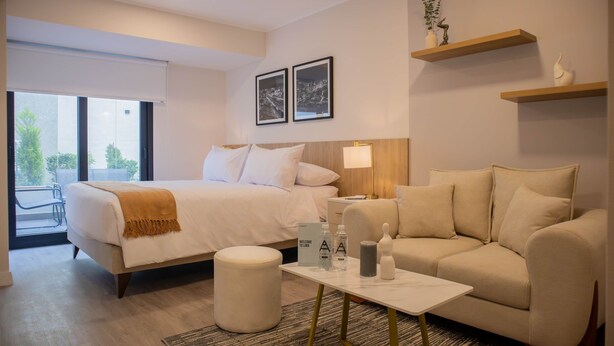 A hotel room with a bed, a sofa, a coffee table, and a small table with a lamp. There are two framed pictures on the wall and a window with a view of trees. - w Spectacular 1BR w Terrace in Miraflores (Lima)