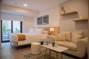 Une chambre d’hôtel avec un lit, un canapé, une table basse et une petite table avec une lampe. Il y a deux tableaux encadrés au mur et une fenêtre donnant sur des arbres.