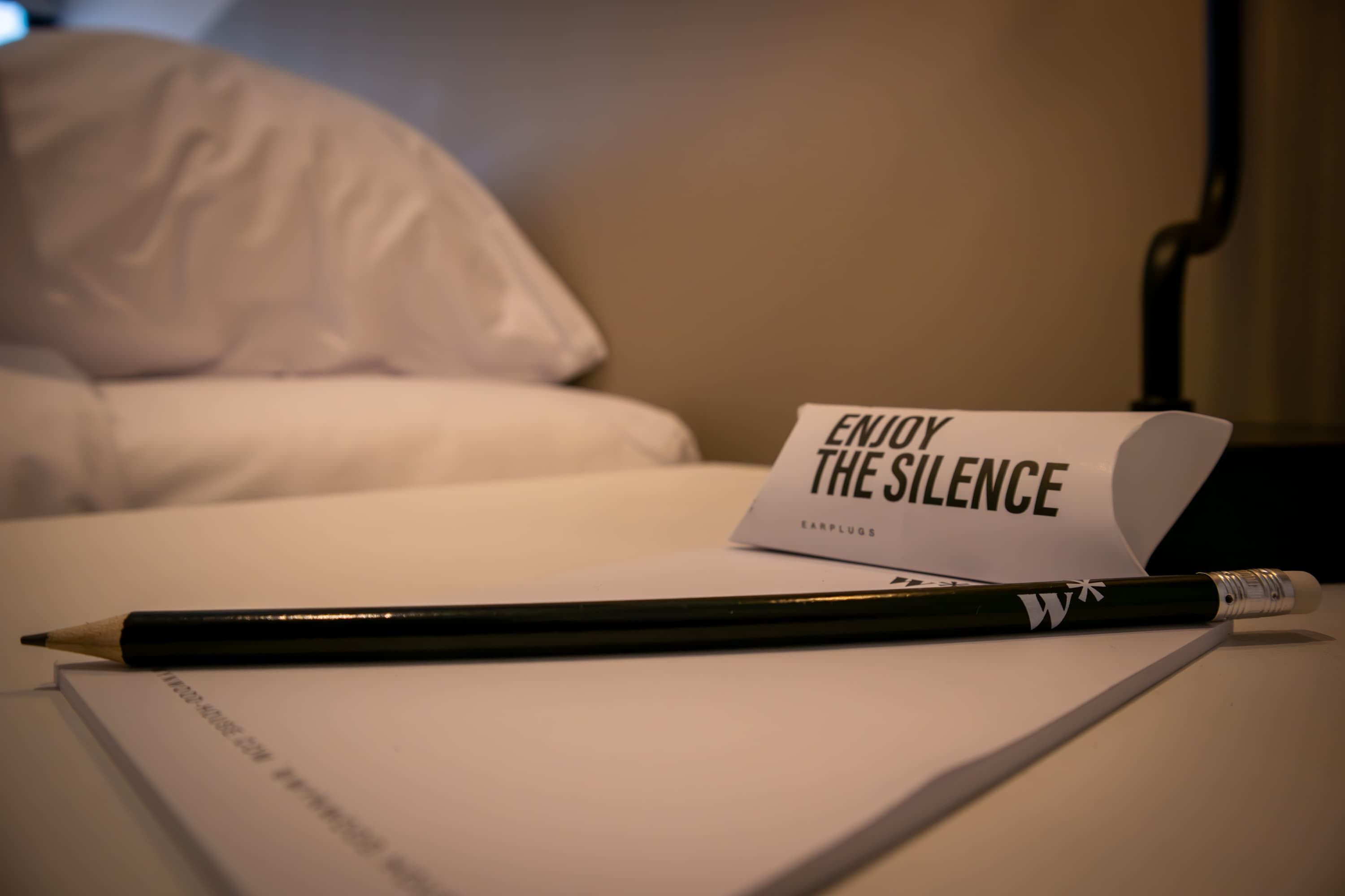 Een bed met witte lakens, een zwarte potlood, en een kaartje met de tekst 'ENJOY THE SILENCE' in een hotelkamer.