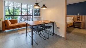 Una habitación moderna con una mesa de comedor de madera, sillas negras, un sofá, una cama con cabecero de madera y ventanas amplias.