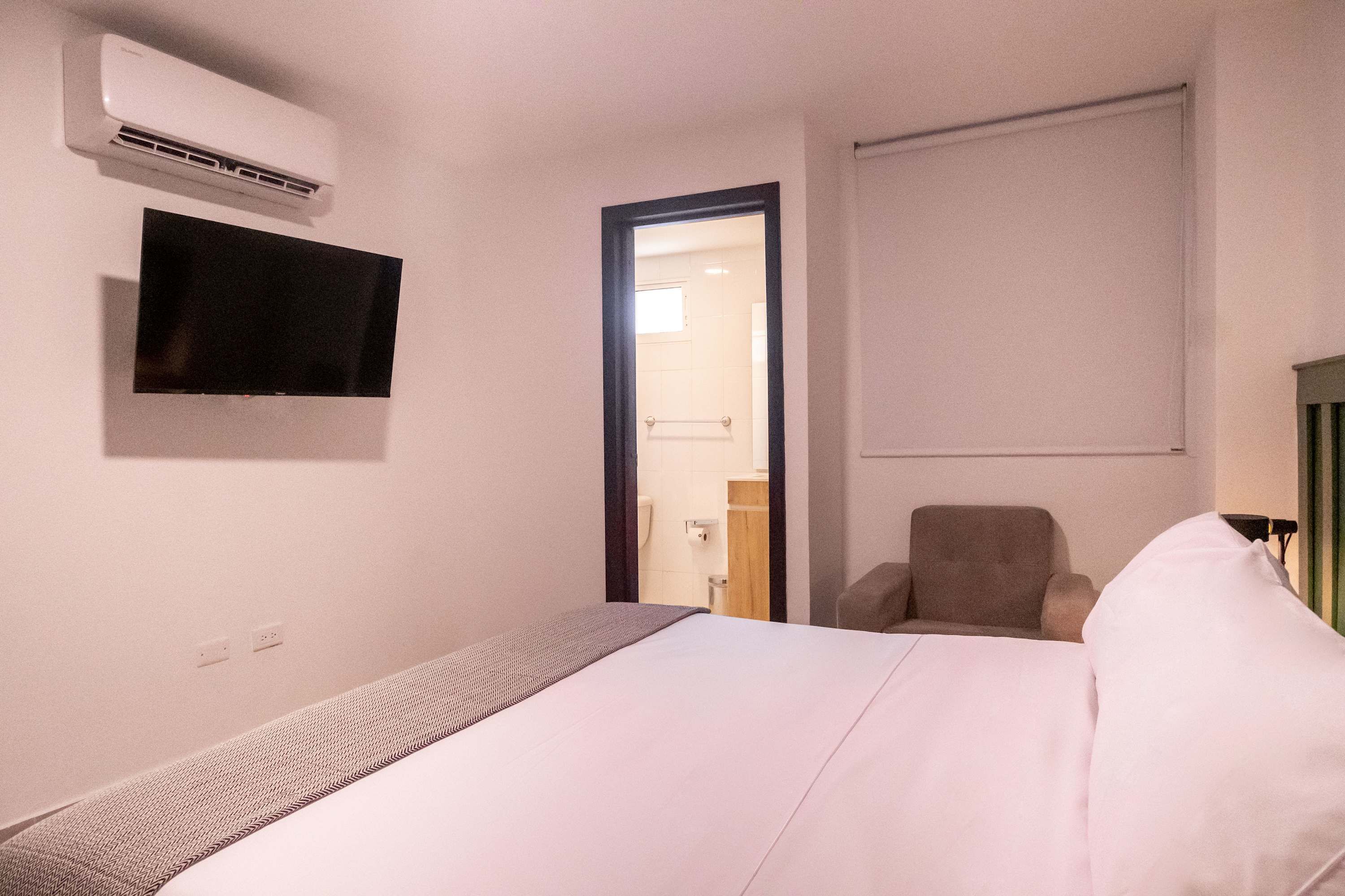 Une chambre d’hôtel comprenant un lit, un téléviseur fixé au mur, un climatiseur, une chaise et une porte donnant sur une salle de bain.