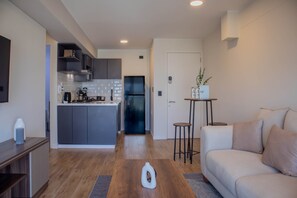 Een moderne keuken met grijze kasten, een centraal eiland en een inbouwoven. In de woonkamer staat een witte bank en een kleine tafel met een plant.