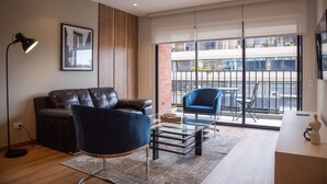 A modern living room with a leather sofa, a glass coffee table, and a blue armchair. A framed picture hangs on the wall, and there is a large window with a view of the outside.