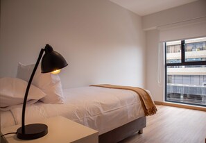 A bedroom with a bed, a bedside table with a lamp, a window with a view of buildings, and a balcony.