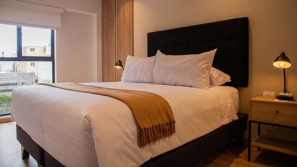 A neatly made bed with white linens and a brown throw, a bedside table with a lamp, and a window with a view of buildings outside.