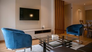 A modern living room with a flat-screen TV mounted on the wall, a glass coffee table, two blue armchairs, and a leather sofa.