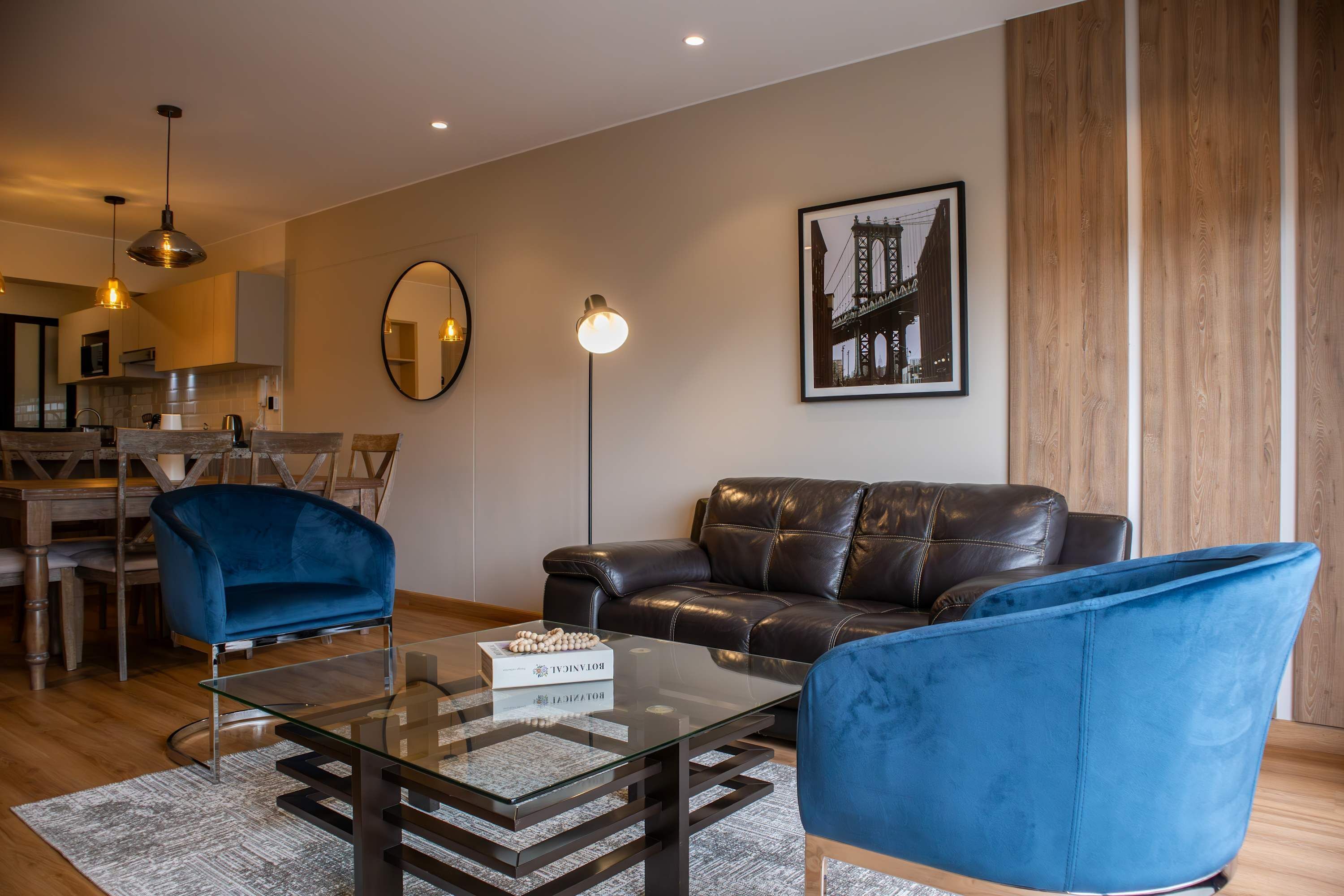 A modern living room with a glass coffee table, two blue armchairs, a leather sofa, a dining area with a wooden table and chairs, and a kitchen in the background.