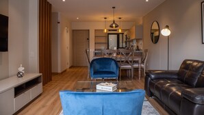 A modern living room with a glass coffee table, a blue armchair, a leather sofa, and a dining area with wooden furniture.