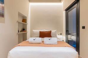 Una cama bien hecha con sábanas blancas, dos toallas con un logotipo, una manta marrón y una pequeña estantería con libros y una planta.