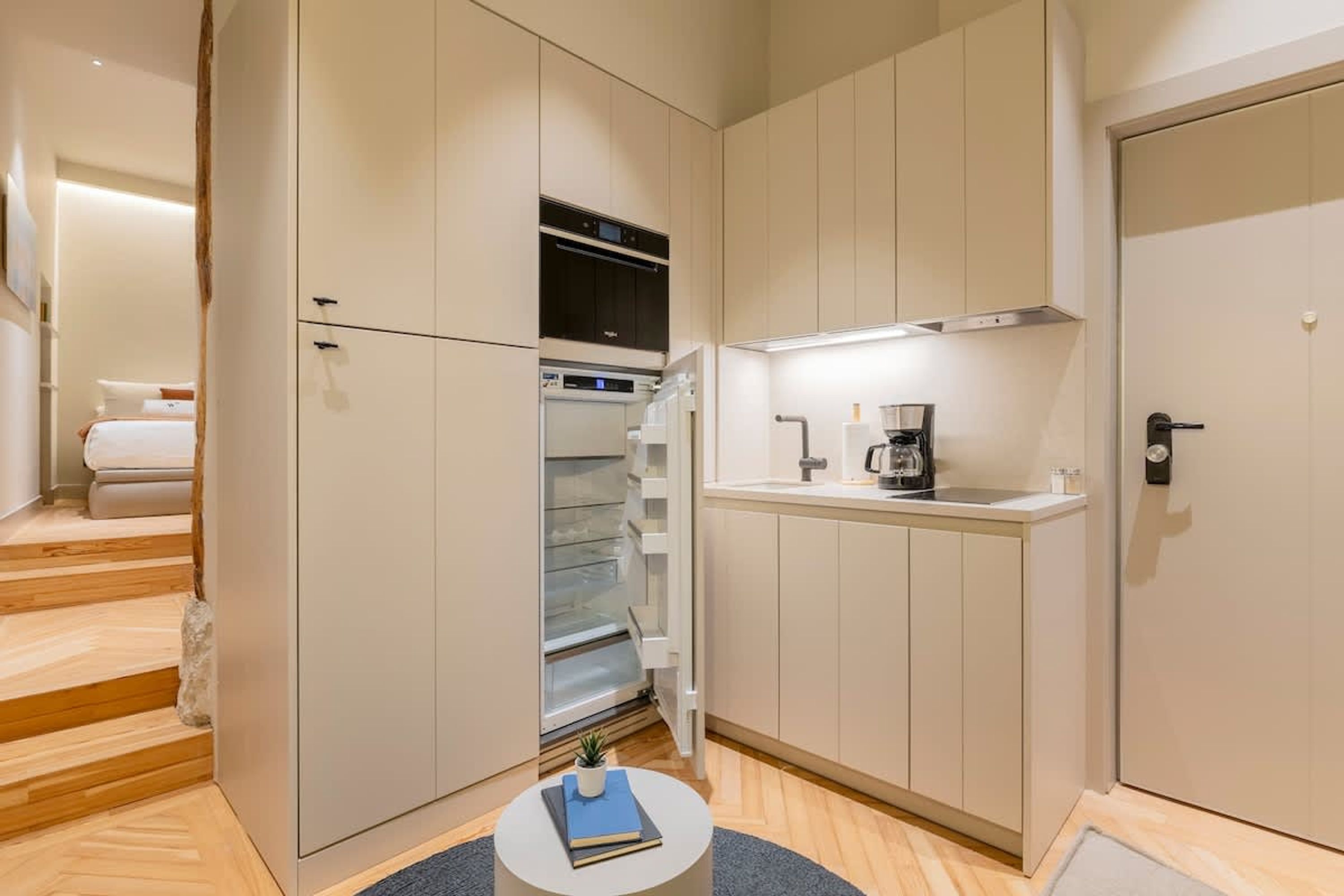 Een compacte hotelkamer met een kitchenette, een klein rond tafeltje met een boek en een bed met een wit hoofdeinde.