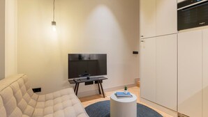 Un saló modern amb un sofà, un televisor de pantalla plana sobre un suport, una taula rodona petita amb un llibre i una planta, i un forn de pared integrat sobre els armaris.