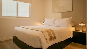 A neatly made bed with a white comforter and a checkered throw blanket, a bedside lamp, and a wall-mounted abstract painting.