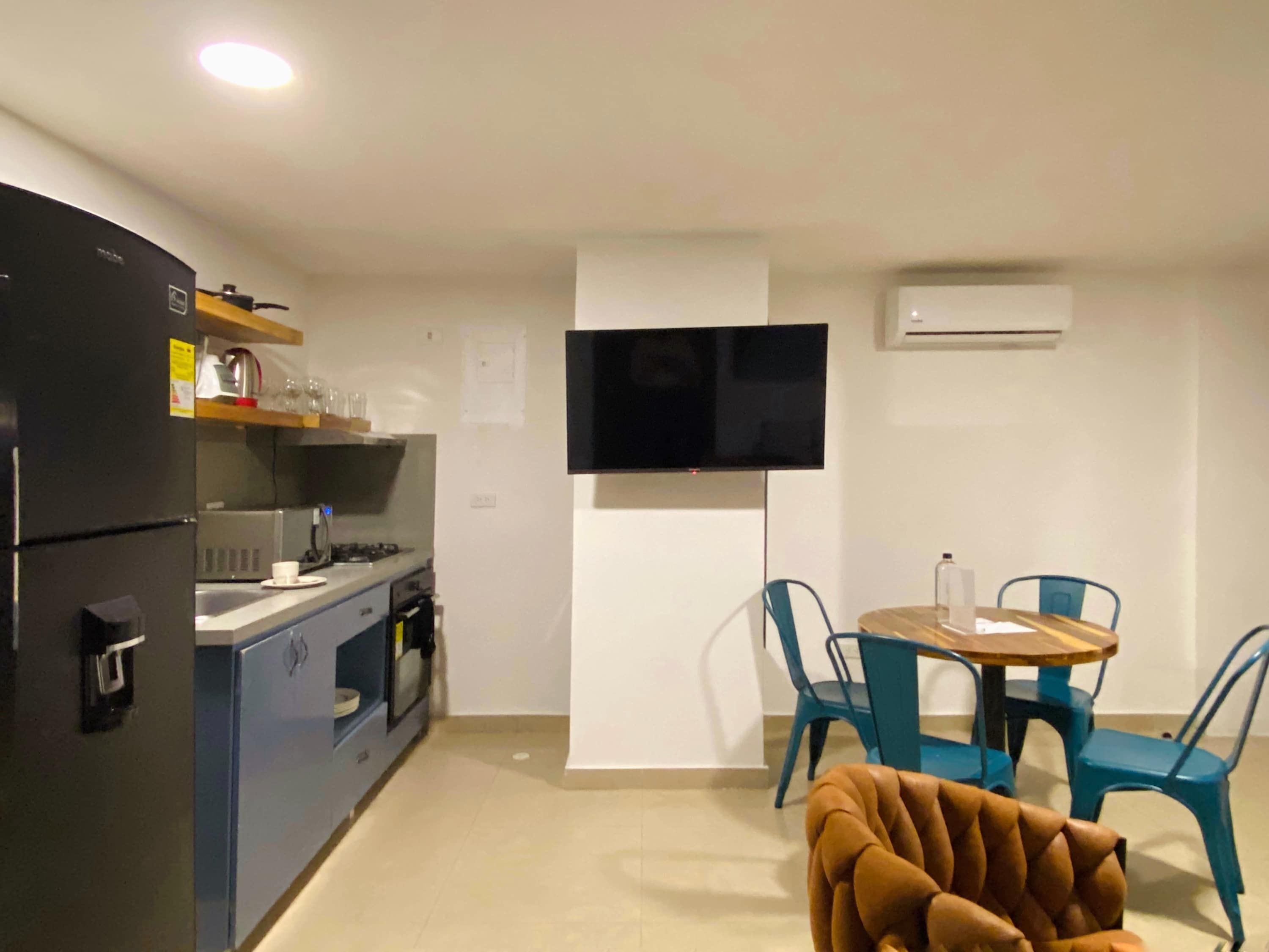 Cocina compacta con refrigerador, estufa y microondas. Cuenta con un comedor con mesa y sillas, y un televisor de pantalla plana montado en la pared.