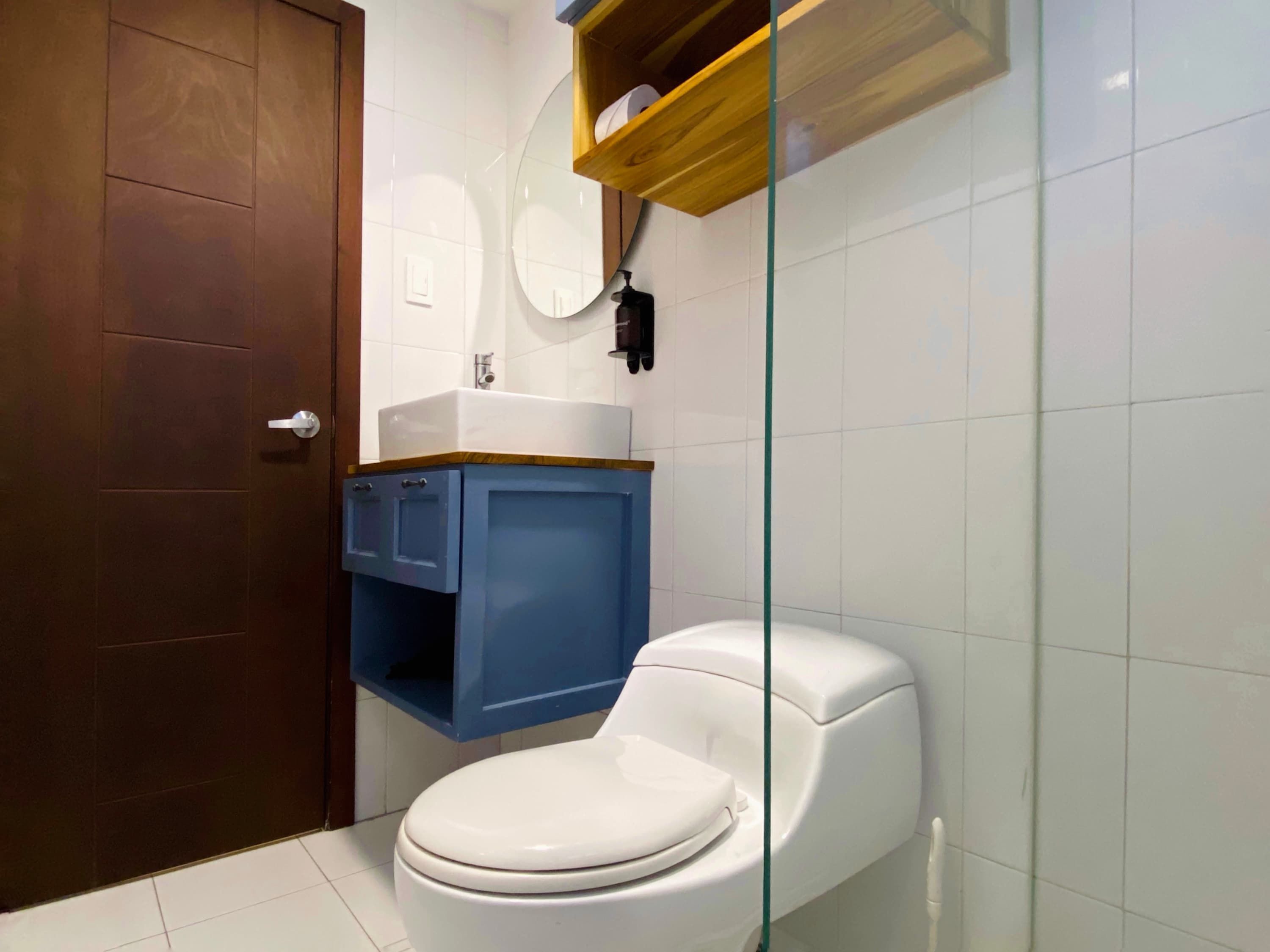 A bathroom with a white toilet, a blue cabinet, a wooden shelf, and a glass shower enclosure.
