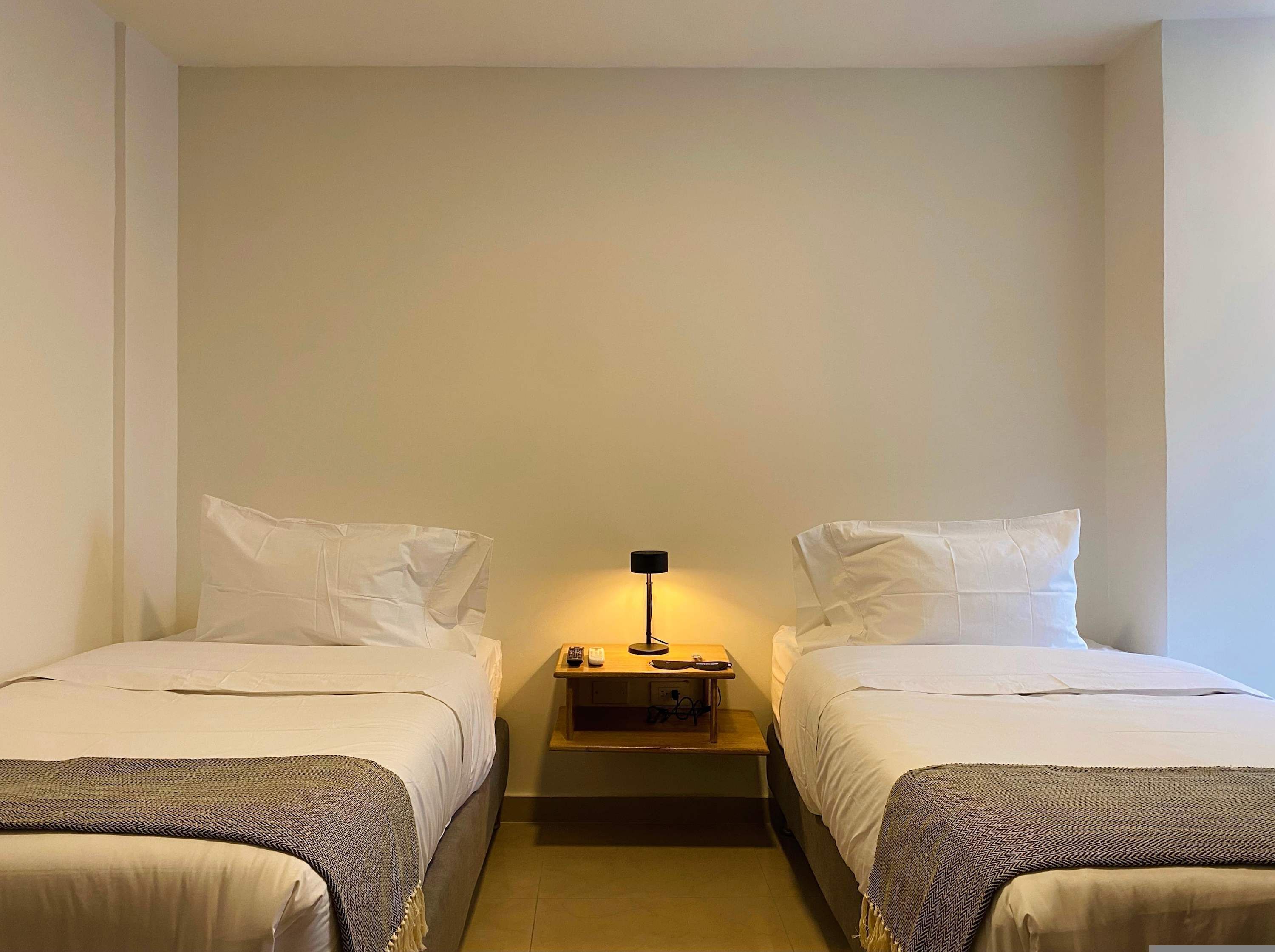 Una habitación de hotel con dos camas individuales, una mesita de noche con lámpara y una pequeña mesita auxiliar de madera.