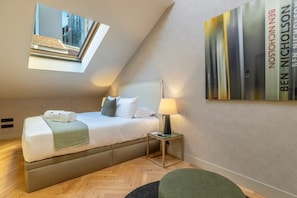 A bedroom with a bed, a bedside table, a lamp, a wall-mounted artwork, and a skylight.