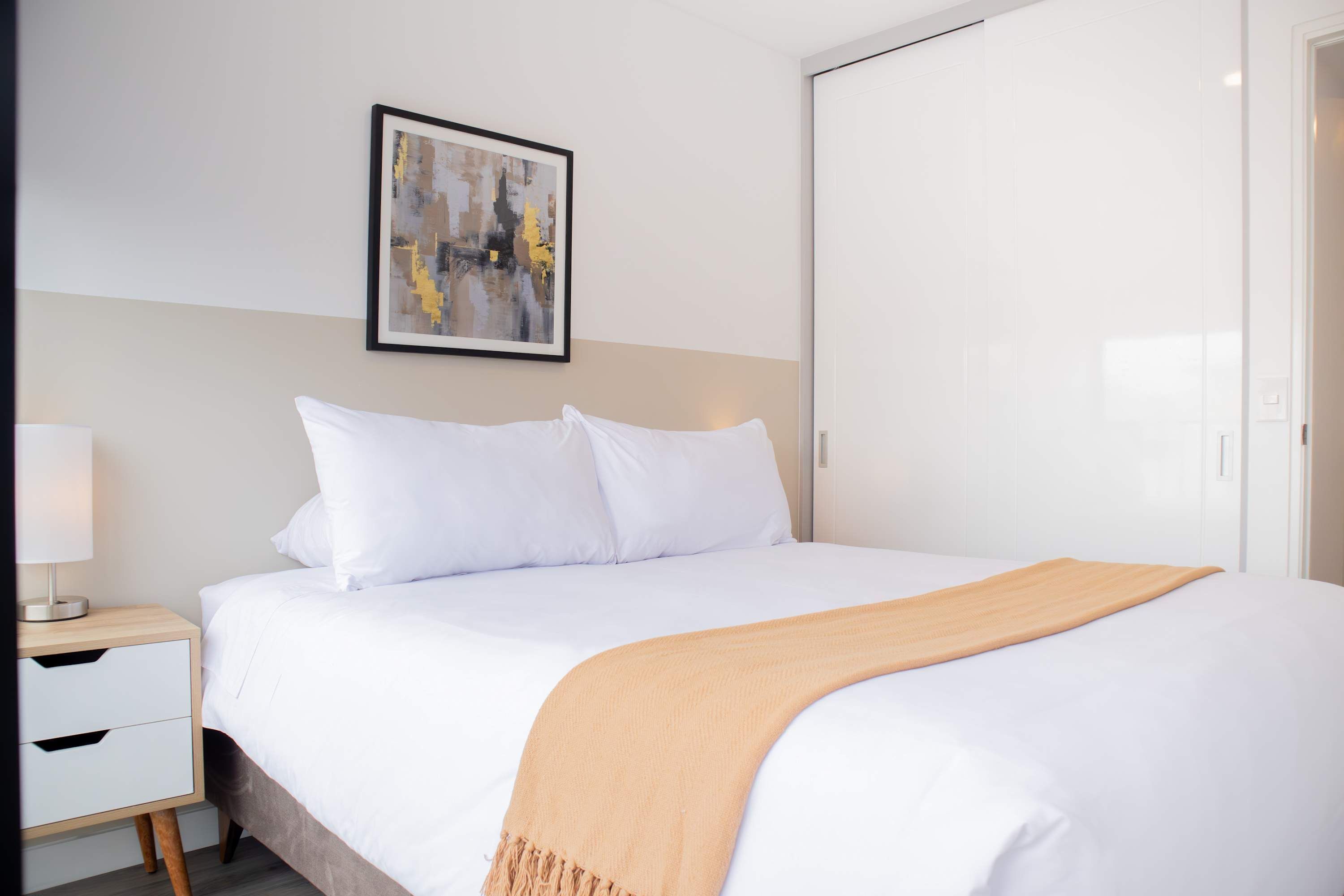 A neatly made bed with white linens and a beige throw blanket, a bedside table with a lamp, and a framed abstract painting on the wall.