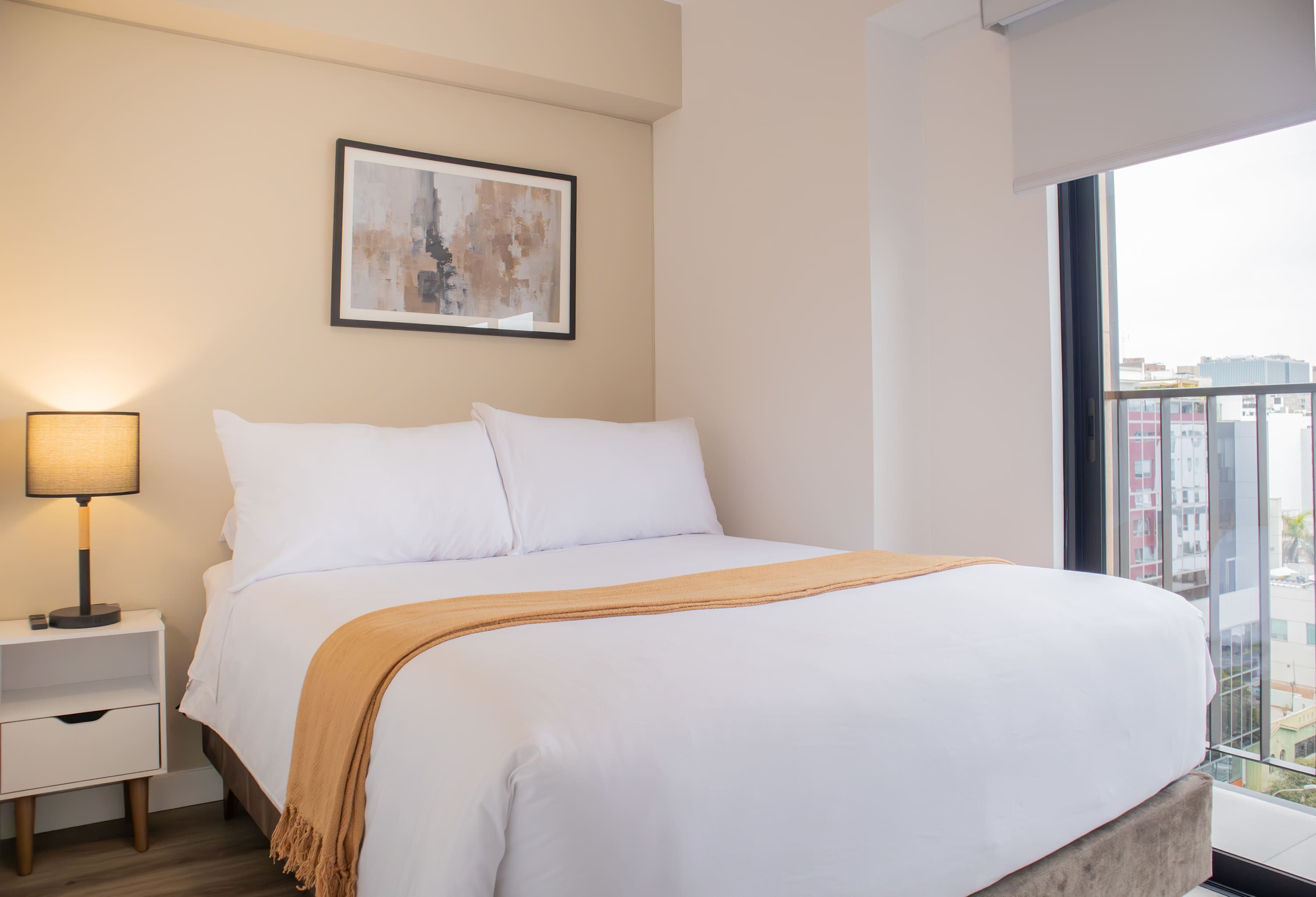 Un cuarto de hotel con cama, mesita de noche, lámpara, un cuadro en la pared y vista de la ciudad a través de un ventanal grande.