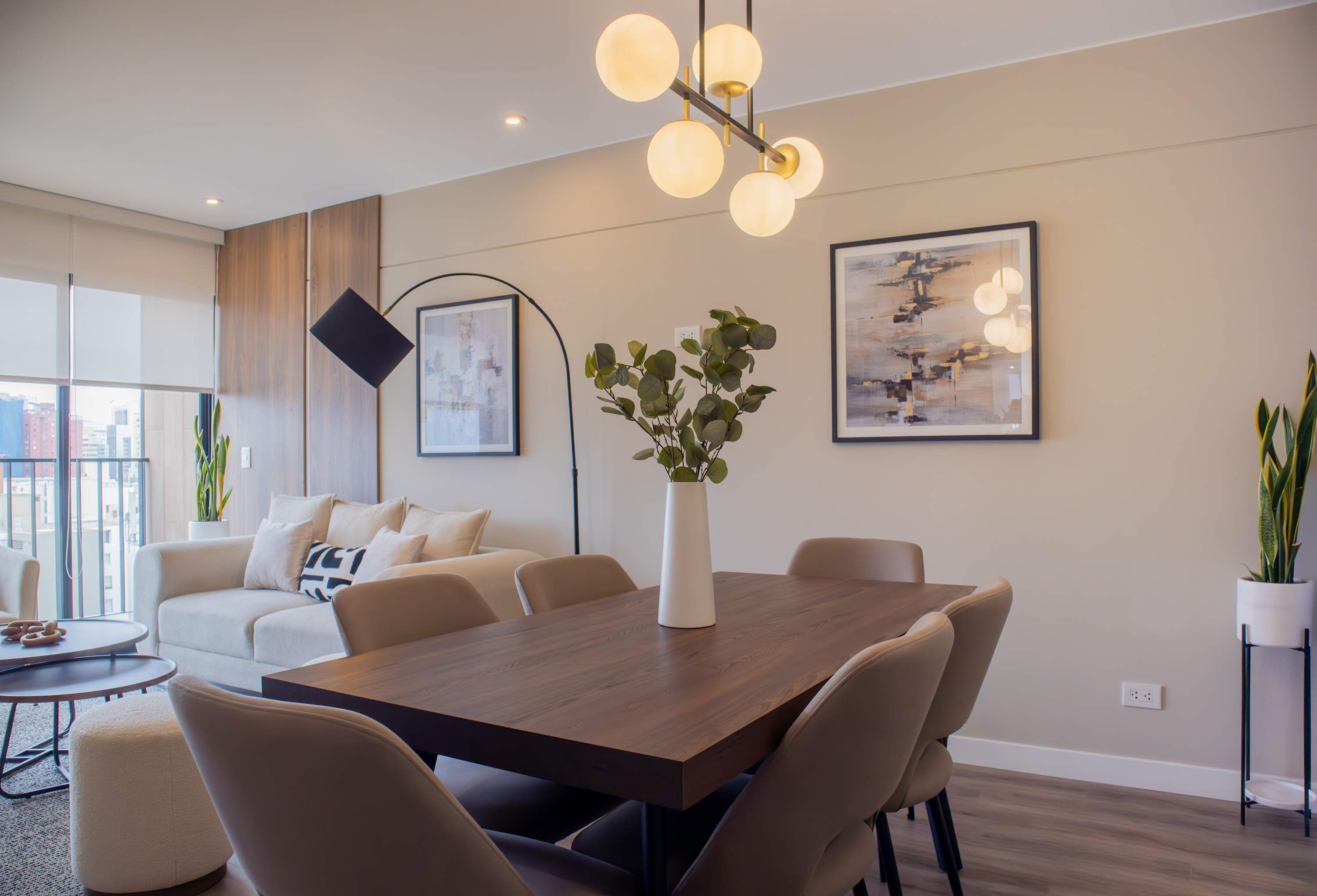 Una sala de estar moderna con una mesa de comedor de madera, sofás color beige y un ventanal con vistas a la ciudad.