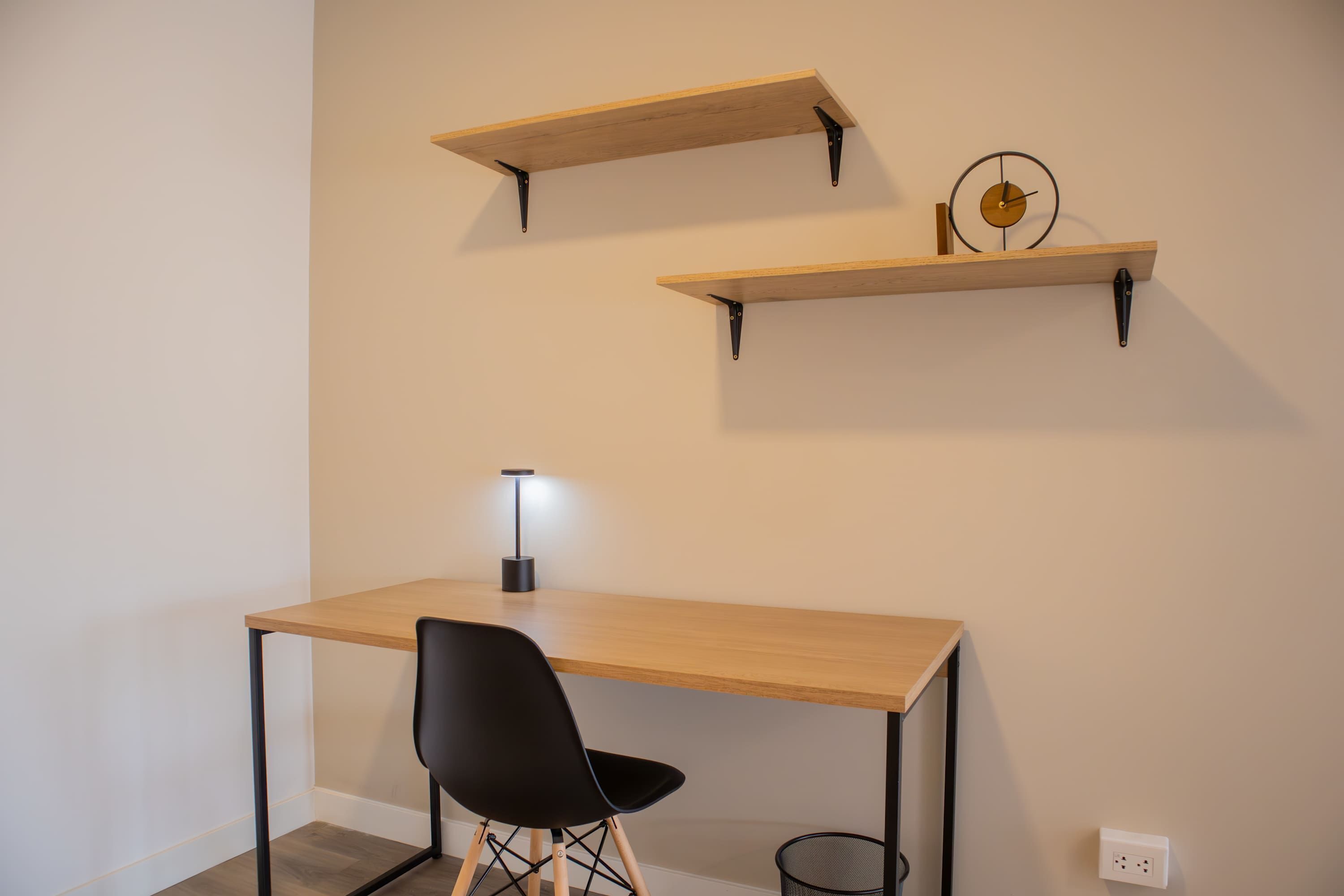 A minimalist room with a wooden desk, a black chair, and two wooden shelves with a clock.