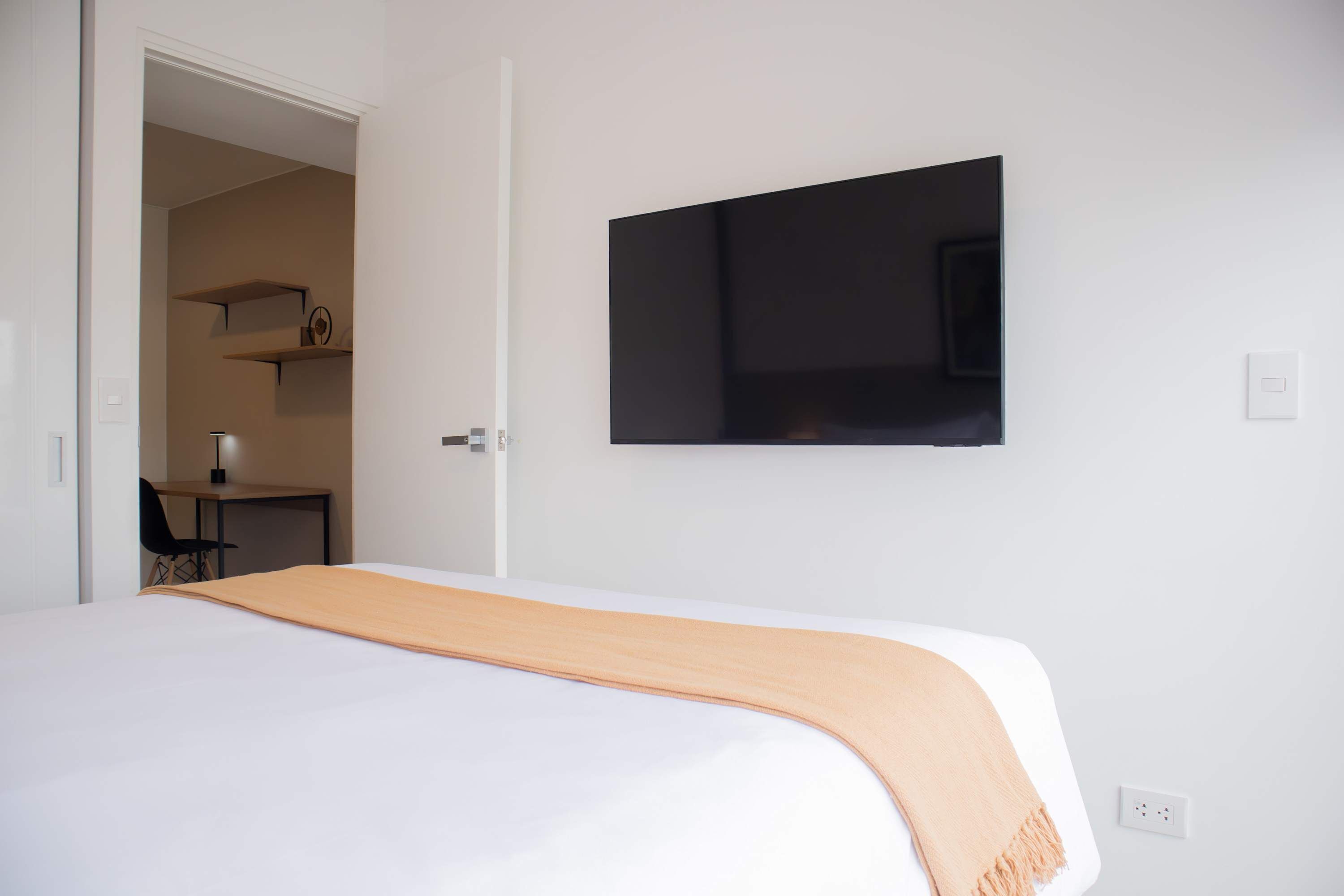 Ein Hotelzimmer mit Bett, Wandfernseher, Schreibtisch mit Stuhl und Ablagefläche mit Gegenständen.