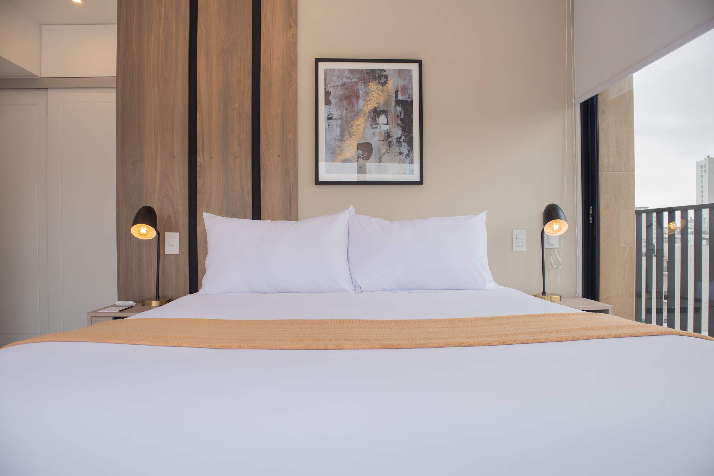 Una habitació d'hotel moderna amb un llit gran, dues làmpades de nit, una obra d'art abstracta emmarcada a la paret i vista a la ciutat a través de la finestra.