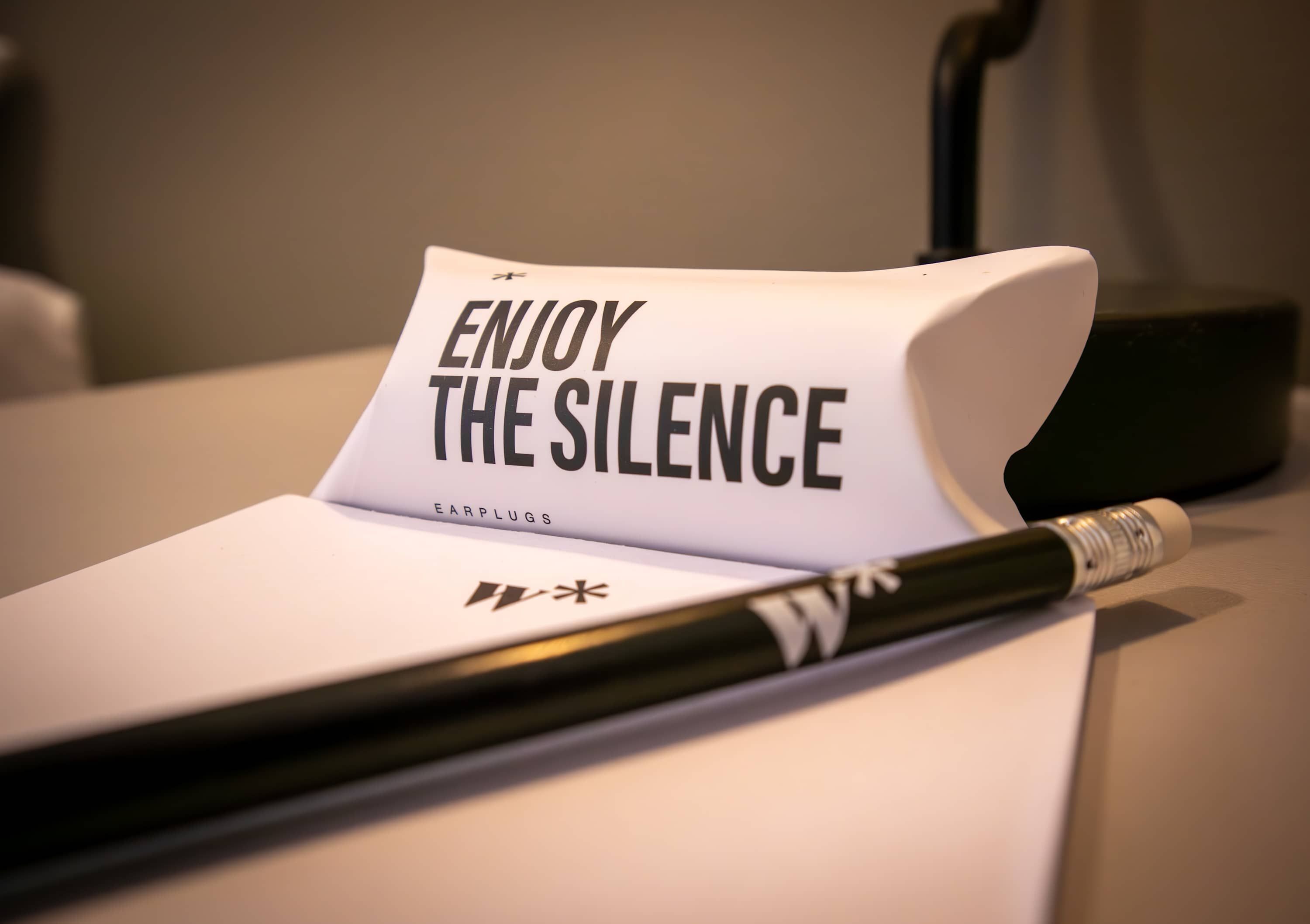 A piece of paper with the text 'ENJOY THE SILENCE' and the word 'EARPLUGS' on it, placed on a surface with a pen lying next to it.