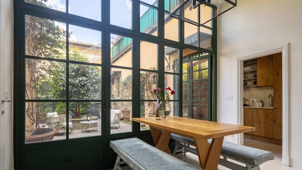 Zona pranzo con tavolo e panche di legno, vista sul giardino attraverso ampie finestre e cucina visibile attraverso una porta aperta.
