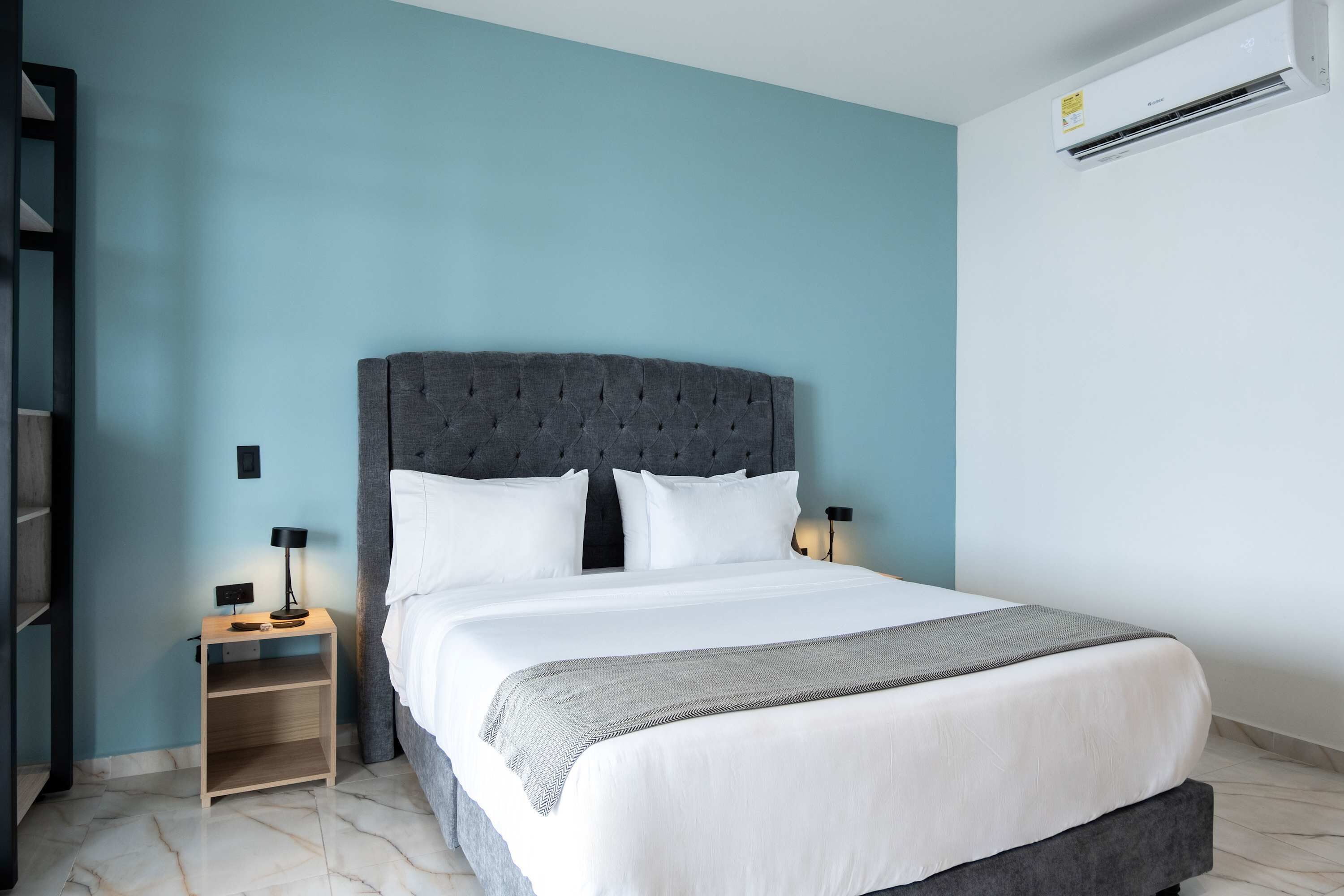 A neatly made bed with a grey upholstered headboard, white bedding, and a grey throw blanket. There are two bedside tables with lamps, and a wall-mounted air conditioner.