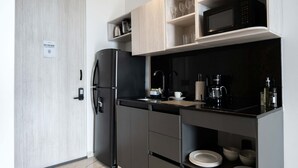 A modern kitchen with a black refrigerator, a microwave, and a coffee maker. There are shelves with cups and glasses, and a door with a 'BYE FOR NOW!' sign.