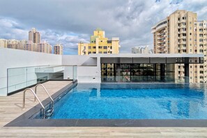 A rooftop swimming pool with a glass railing, surrounded by modern buildings and a cloudy sky.