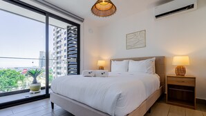 Quarto moderno com uma cama grande, duas mesas de cabeceira com luminárias, uma planta em vaso e vista para a cidade através de uma janela ampla.