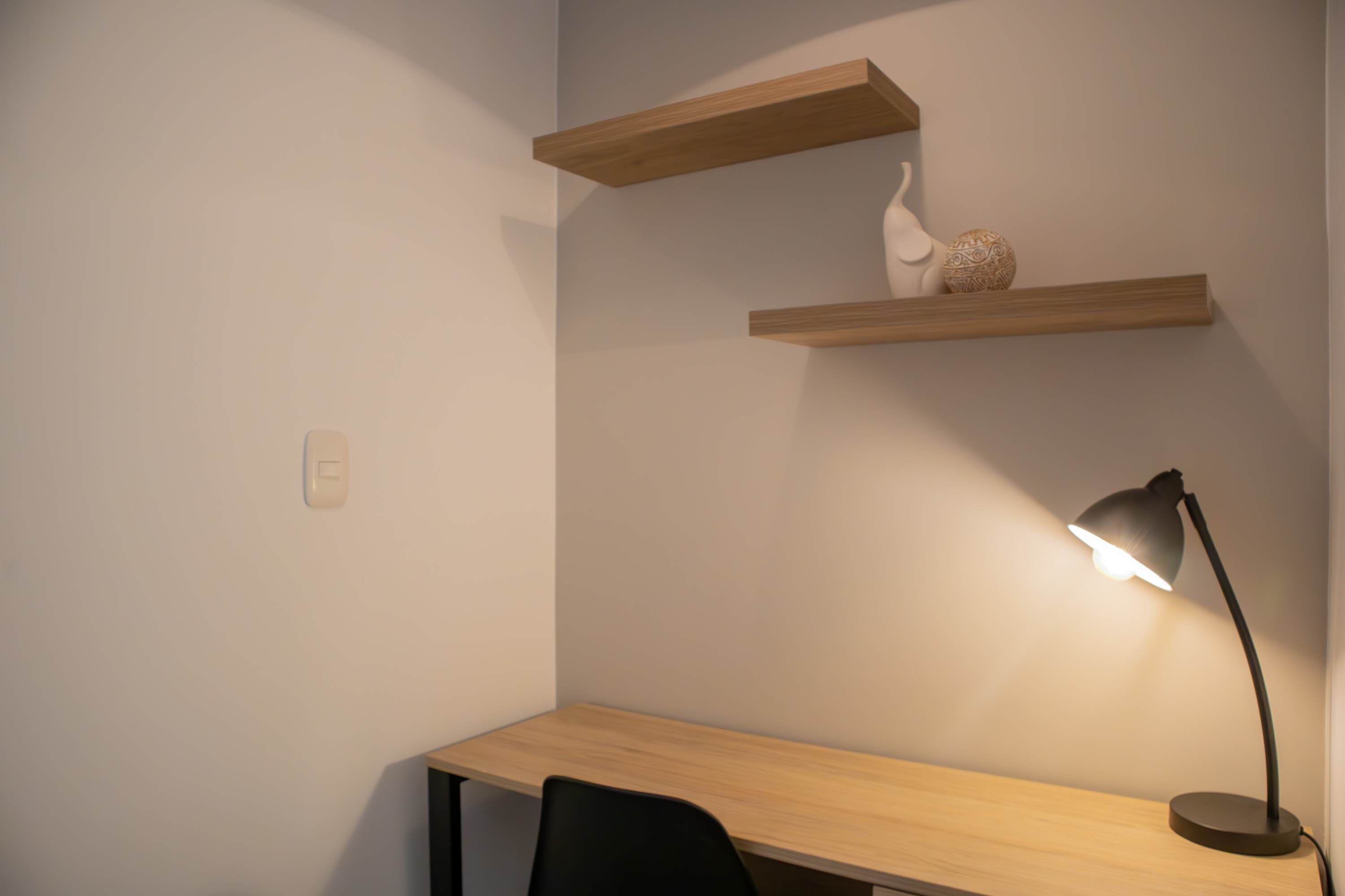 A corner desk with a black chair, a desk lamp, and two wooden shelves with decorative items.