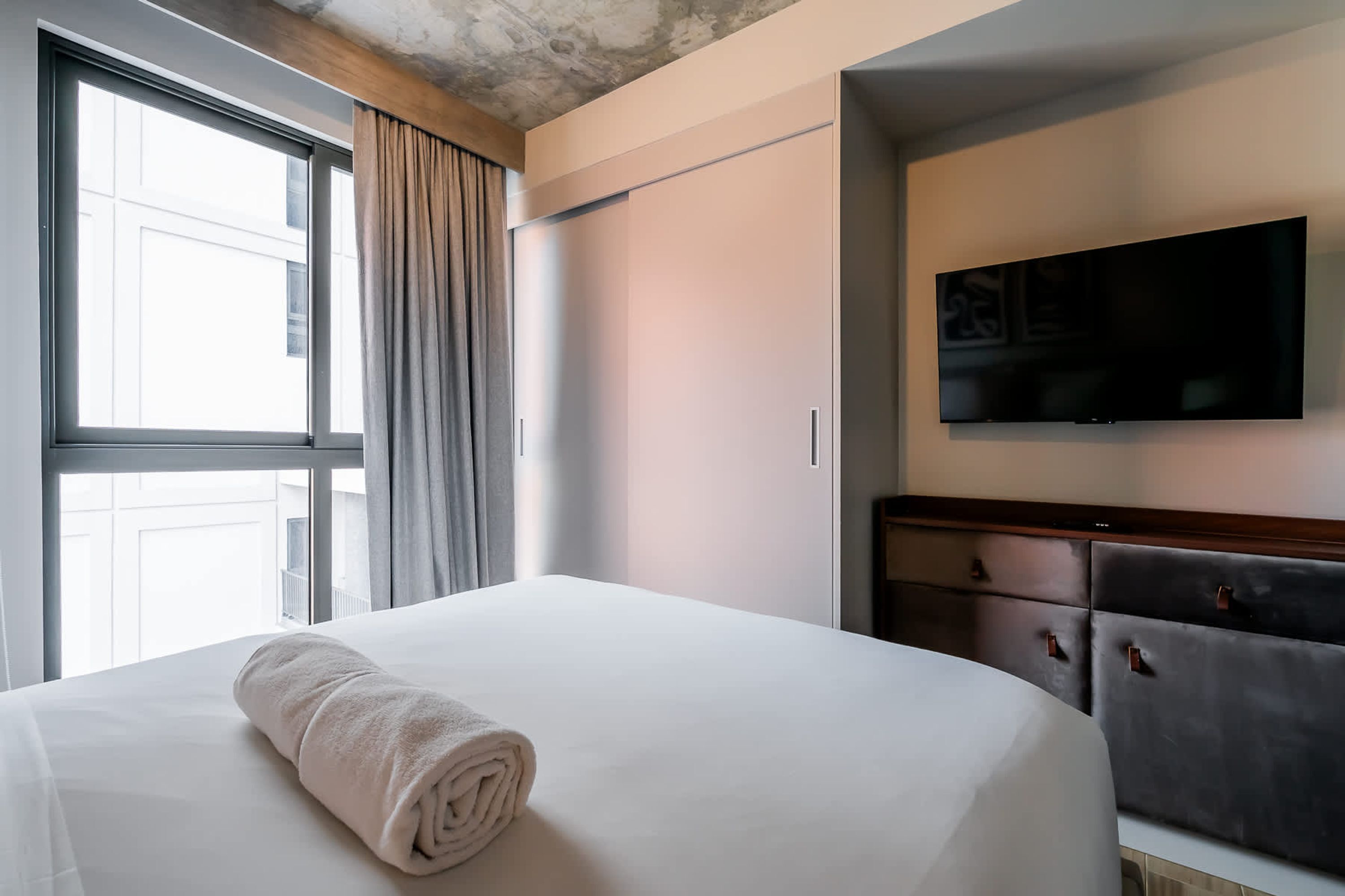A hotel room with a large bed, a flat-screen TV mounted on the wall, and a window with curtains.